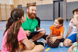 © LIGHTFIELD STUDIOS - A man sits on the floor surrounded by a group of kids, teaching with enthusiasm in a colorful classroom setting.