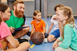 © LIGHTFIELD STUDIOS - A diverse group of children sit attentively around a basketball as their male teacher instructs them in a bright, lively classroom setting.