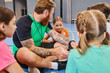 © LIGHTFIELD STUDIOS - A man sitting on the floor surrounded by a group of diverse children, engaging in lively instruction in a bright classroom setting.