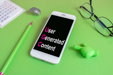 UGC user generated content symbol. Concept words UGC user generated content on the mobile phone screen. Beautiful green background. Glasses, pen, coins. 