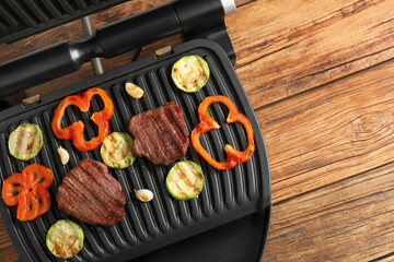 Wall Mural - Electric grill with tasty meat steaks and vegetables on wooden table, top view. Space for text