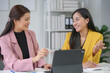 © amnaj - Two young businesswomen are sitting at their desk in the office, smiling and discussing while working on a new project using a tablet