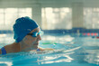 © Victor Bertrand - An elderly swimmer, wearing a blue swim cap and goggles, glides through a sunlit indoor pool with focused determination and grace.