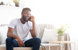 © Prostock-studio - African American man with a beard sits on a white couch in a living room, smiling and laughing while talking on the phone. He is wearing a white t-shirt and blue jeans, copy space