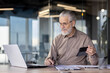 © Liubomir - Senior businessman working on a laptop and holding a tablet in a modern office setting. The professional atmosphere showcases productivity, experience, and the use of technology business operations.