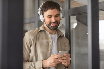 Aufkleber - A man in a tan shirt and white t-shirt is standing in an office setting. He is wearing headphones and looking down at his smartphone. He appears to be listening to something on his phone.