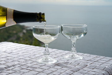 Wall Mural - Pouring of cold sparkling wine, cava or champagne in two coupe glasses on table with sea water view