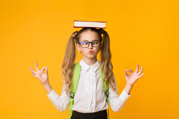 Wall Mural - Back To School Concept. Cute Nerdy Schoolgirl Meditating With Book On Head On Yellow Studio Background. Free Space