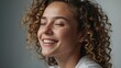© Adam - A joyful young woman with curly hair smiling brightly with closed eyes, radiating happiness against a plain background.
