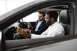 © sofiko14 - Car dealership, sales, and customer service. Caucasian car dealer sit inside vehicle with African American buyer, discussing car features. Modern business interaction focusing on buying new car.