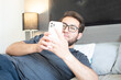 © Diego D - Hombre apuesto con anteojos relajado en su habitación. Joven apuesto usando su teléfono celular