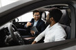 © sofiko14 - Caucasian car dealer sit inside vehicle with African American buyer discussing purchase details with friendly demeanor
