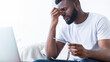 © Prostock-studio - African American man is seated in front of a laptop, his hand pressed against his eye, indicating discomfort and eye strain. Struggle of eye fatigue associated with prolonged screen time.