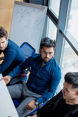 Wall Mural - Creative multiethnic business team in the modern conference room discuss work results siiting at the table against big window background. Focus on handsome Indian guy in dark formal shirt.