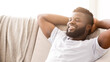 © Prostock-studio - Black man relaxing on a couch. He is wearing a white t-shirt and has his arms behind his head. His eyes are closed and he is smiling. He appears to be enjoying a moment of peace and relaxation.