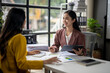 © Wasana - Two women are sitting at a desk, one of them holding a tablet