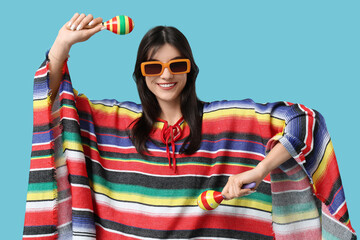 Wall Mural - Young woman in colorful poncho with maracas on blue background. Cinco de Mayo celebration