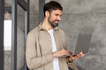 Aufkleber - A man with a beard is standing near a glass door and working on his laptop. He is wearing a light brown button-down shirt over a white t-shirt. His expression is focused as he types on the keyboard.
