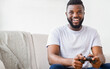© Prostock-studio - Black young man with a beard sits on a couch while playing a video game. He is smiling and holding a game controller. The background is a simple white wall, copy space