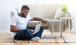 © Prostock-studio - A young black man sits on the floor of his living room, using a laptop computer. He is sitting on a rug in front of a couch, with a plant on a side table
