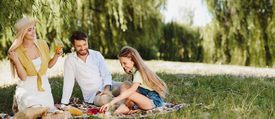 Wall Mural - Happy parents picnicking with their cute daughter, sitting on blanket in park