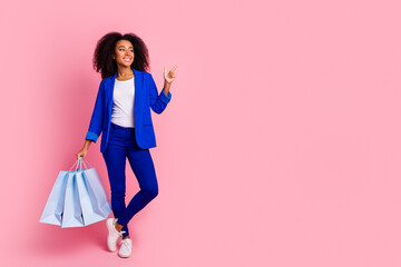 Poster - Full length photo of cheerful woman wear blue jacket hold shopping bags look directing empty space isolated on pink color background