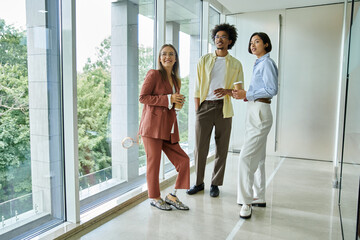 Three colleagues stand near a large window in a modern office, engaged in a friendly conversation.