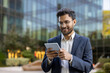 © Liubomir - Smiling businessman using digital tablet outside modern glass office building. Professional wearing suit and glasses. Concept of technology, success, and business communication in urban environment.