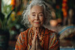 © DKPhoto - An elderly woman peacefully meditates with her hands in the Namaste position, surrounded by colorful, serene decor, embodying spirituality, wisdom, and inner peace.