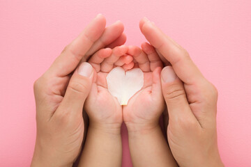 Heart shape of beautiful fresh white peony petal on baby girl and young adult mother opened palms on pink table background. Pastel color. Closeup. Point of view shot. Love concept. Top down view.