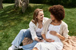 © Bliss - Two women, one pregnant, share a tender moment while sitting on a blanket in a grassy park.