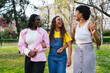 © Koldo_Studio - Three young African women laughing and walking arm-in-arm in a vibrant green park.