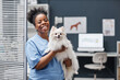 © Seventyfour - Medium shot portrait of trustworthy female vet expert of Black ethnicity smiling looking at camera holding happy white pomeranian dog after vaccination in vet clinic, copy space