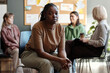 © pressmaster - Young depressed woman in t-shirt and pants sitting on chair in front of group of patients and female psychologist having discussion
