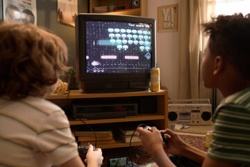 Back view of two kids absorbed in playing a retro video game on CRT TV, sitting on a cozy living room carpeted floor with snacks around