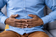 © Liubomir - Close-up of man's hands clutching stomach indicating abdominal pain and discomfort. Concept of digestive issues, stomach ache, and health problems. Man in blue shirt and brown pants sitting on couch.