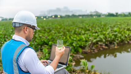 Biologist wastewater treatment sit aside of a lake while looking at the wastewater in a plastic glass on his hand and check the quality of the water sample and contaminants, laptop, analysis, 