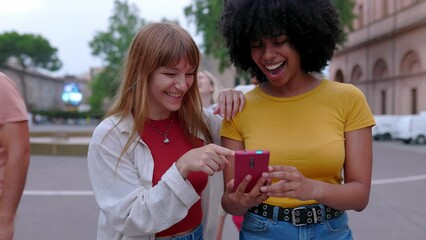 Wall Mural - Two happy young diverse women having fun watching social media content on mobile phone app while walking with some friends at city street. Friendship and technology concept. Slow motion video.