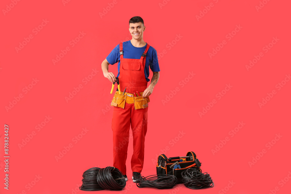 Young male electrician with tool bag and cables on red background