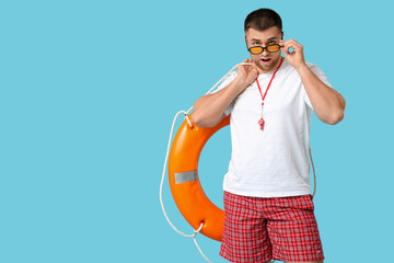 Wall Mural - Surprised young lifeguard in sunglasses with lifebuoy on blue background