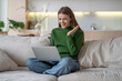 © DimaBerlin - Pleasantly surprised girl smiling looks at screen laptop, sits on comfortable couch, chatting online, booking. Delight woman with computer make internet shopping, find sale, glad by discount at home.