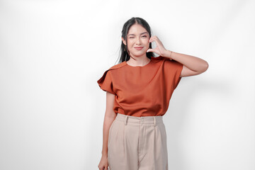 Wall Mural - Cute young Asian woman in a brown shirt making a heart gesture on her cheek, isolated over white background.