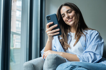 Happy pleasant millennial woman relaxing on comfortable couch, holding smartphone in hands. Smiling young lady chatting in social networks, watching funny videos, using mobile applications at home.