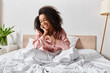 © LIGHTFIELD STUDIOS - A curly African American woman in pajamas sits calmly on a bed with white sheets in a serene bedroom in the morning.