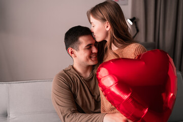 Concept of Happy Valentine's Day. Young couple in love holding a heart-shaped balloon sitting on the sofa at home and kissing. Romantic evening.