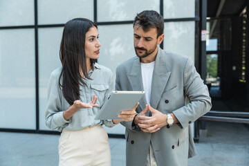 Two coworkers using a digital tablet and talking while standing outside. Modern business colleagues chatting together in front of office building on their way to work. Businesspeople collaboration