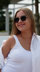 Wall Mural - Blonde girl sitting on deckchair in white clothes wearing sunglasses. Smiling woman covers her shoulder from sun. Vertical video