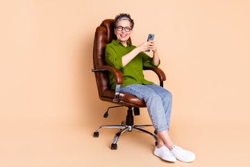 Poster - Full size photo of pretty senior female hold device sit armchair wear trendy green outfit isolated on beige color background