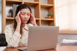 © Ekkasit A Siam - Asian female with long hair feels stressed, hands on temples, in front of laptop at a wooden desk, papers stacked beside, in room with shelves. Office worker experiences tension, massaging forehead