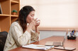 © Ekkasit A Siam - Asian female office worker taking a break, sipping from a cup at a wooden desk. Casual attire in a modern office setting with documents and a tablet on the desk. Reflective and calm expression.